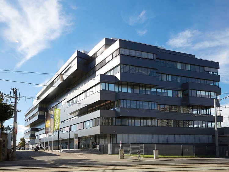 Photo: multi-storey office block with a dark grey aluminium-glass façade consisting in part of projecting elements; colourful flags in front of the entrance; tram rails in the foreground
