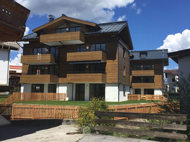 Visualisation: three-storey residential building in a style inspired by local rural architecture, with balconies, wooden façade and a gabled roof, surrounded by a wooden fence and green spaces